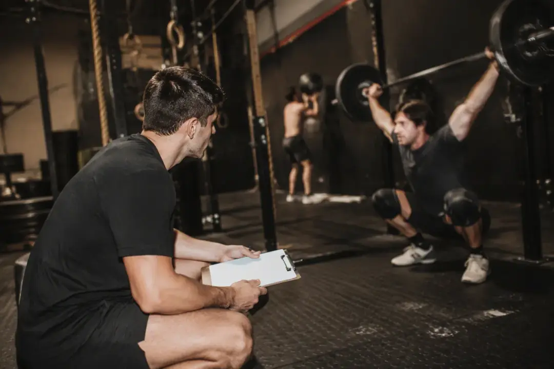 Un coach con una planilla observa a un atleta en una sentadilla con barra